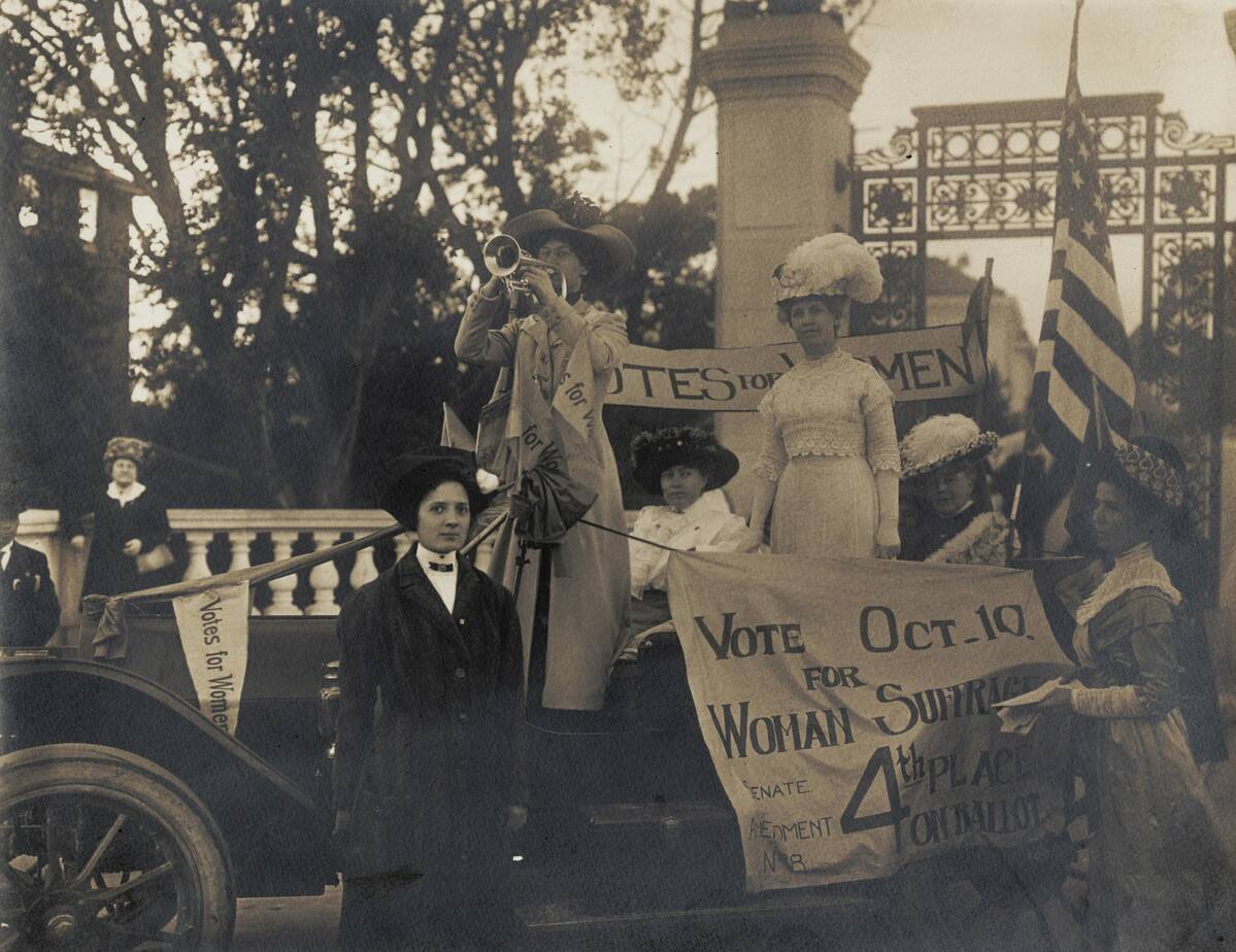 View of Margaret A. Haley wearing a plumed hat and standing in a car n a women's suffrage parade, other women stand nearby, one plays a trumpet